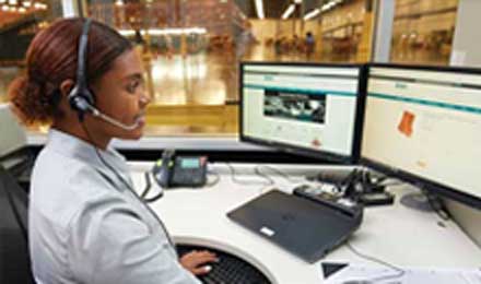 Set in a warehouse or distribution center, a smiling worker with a headset sits at a large desk and works at a computer displaying the Airgas.com homepage.