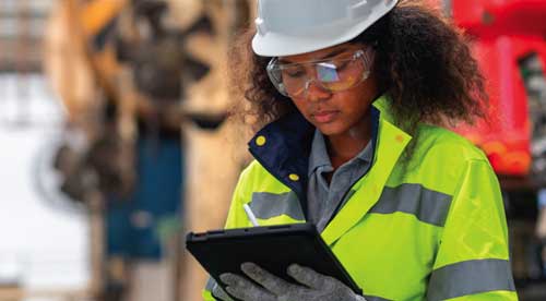 Set in a warehouse or industrial plant or facility, a man wearing personal protective equipment (PPE), including an Airgas branded hardhat, high-visibility safety vest and safety gloves, holds a clipboard.