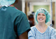 Set in a hospital hallway, a woman wearing scrubs and a disposable hair net smiles at a healthcare worker wearing scrubs with his back to the camera.