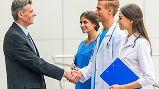 Set in a hospital, a doctor, surrounded by another doctor and a nurse, happily shakes hands with a man in a suit.
