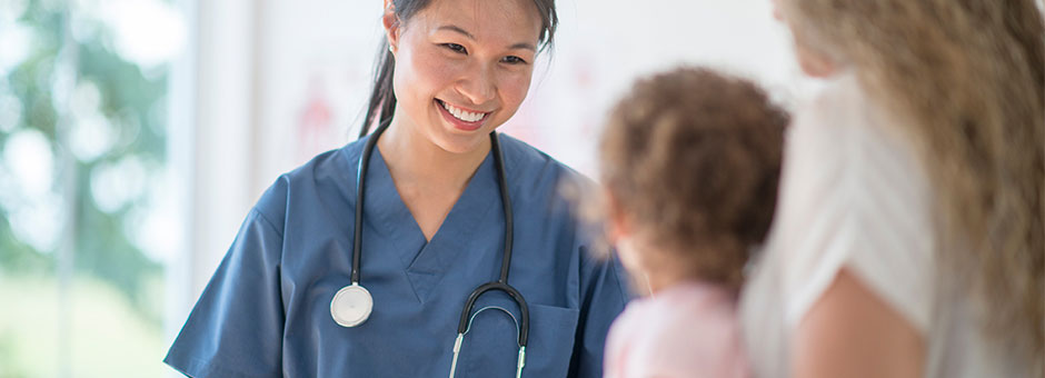 A nurse in scrubs smiles at a mother and her young daughter.