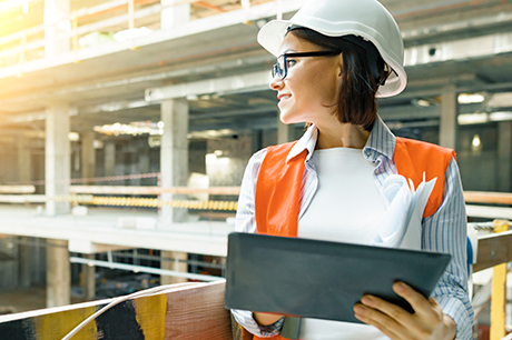 Set on a commercial or industrial construction site, a woman wearing personal protective equipment (PPE), including a hardhat, safety glasses and a high-visibility safety vest, holds a tablet computer while looking down on the site from an upper floor.