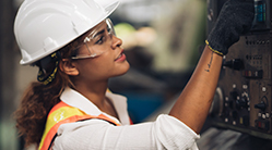 A woman wearing personal protective equipment (PPE), including a hardhat, safety glasses, gloves and a high-visibility safety vest, works on equipment