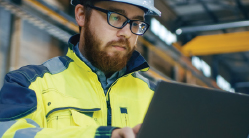 A man wearing personal protective equipment (PPE) in an industrial plant, warehouse or facility, types on a laptop computer.