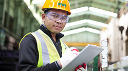 Set in a warehouse or industrial plant or facility, a man wearing personal protective equipment (PPE), including an Airgas branded hardhat, high-visibility safety vest and safety gloves, holds a clipboard.