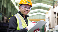 Set in a warehouse or industrial plant or facility, a man wearing personal protective equipment (PPE), including an Airgas branded hardhat, high-visibility safety vest and safety gloves, holds a clipboard.