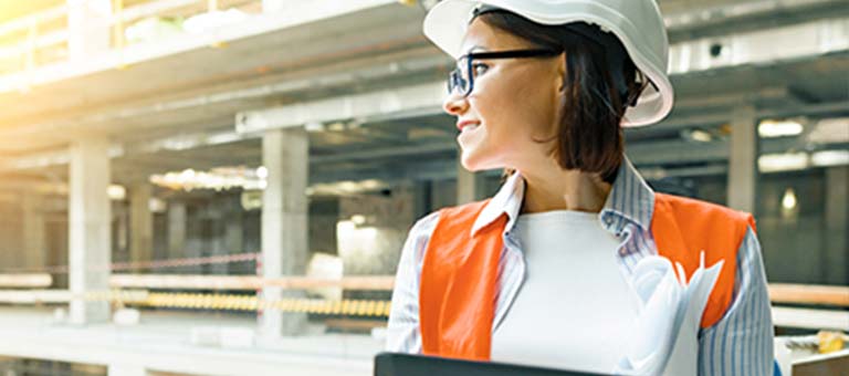 Set on a commercial or industrial construction site, a woman wearing personal protective equipment (PPE), including a hardhat, safety glasses and a high-visibility safety vest, holds a tablet computer while looking down on the site from an upper floor.