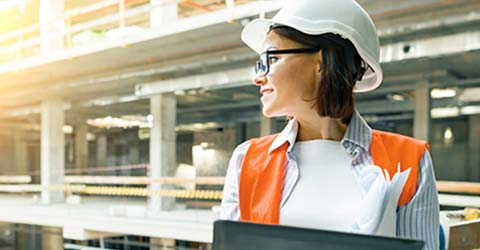 Set on a commercial or industrial construction site, a woman wearing personal protective equipment (PPE), including a hardhat, safety glasses and a high-visibility safety vest, holds a tablet computer while looking down on the site from an upper floor.