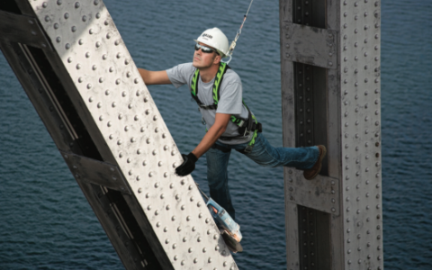 A worker wearing a Honeywell Miller fall-protection harness scales a beam.