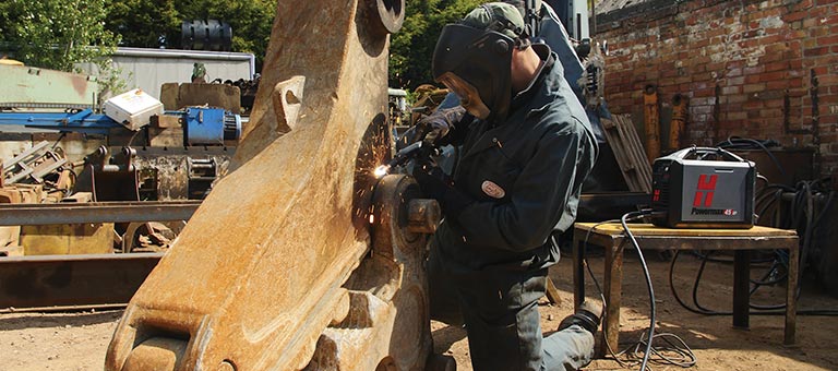While kneeling, a man wearing welding safety helmet, welding jacket and welding gloves, uses a Hypertherm Powermax plasma-cutting system on a large machine part outside in the workyard.