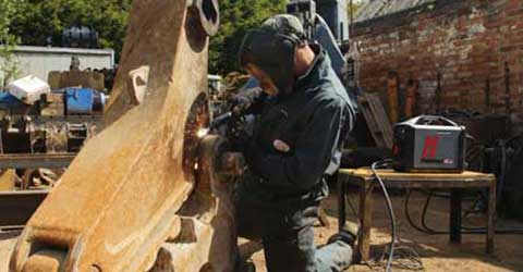 While kneeling, a man wearing welding safety helmet, welding jacket and welding gloves, uses a Hypertherm Powermax plasma-cutting system on a large machine part outside in the workyard.