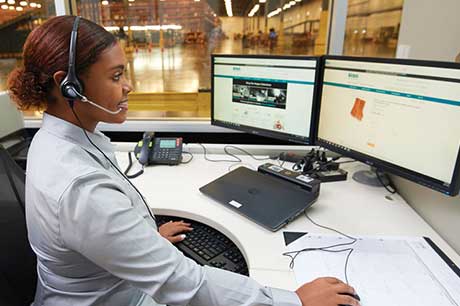 Set in a warehouse or distribution center, a smiling worker with a headset sits at a large desk and works at a computer displaying the Airgas.com homepage.