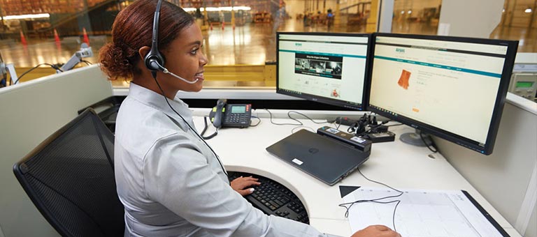 Set in a warehouse or distribution center, a smiling worker with a headset sits at a large desk and works at a computer displaying the Airgas.com homepage.