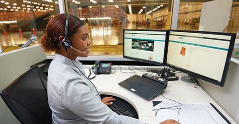 Set in a warehouse or distribution center, a smiling worker with a headset sits at a large desk and works at a computer displaying the Airgas.com homepage.