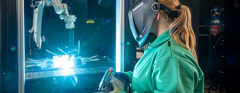 A female welding operator wearing a RADNOR welding helmet, operates an automated welding machine performing a weld.  