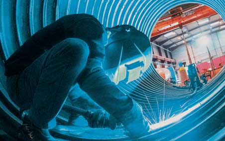 Welder standing inside of a large corrugated metal pipe performing a weld on the interior of the pipe underneath.