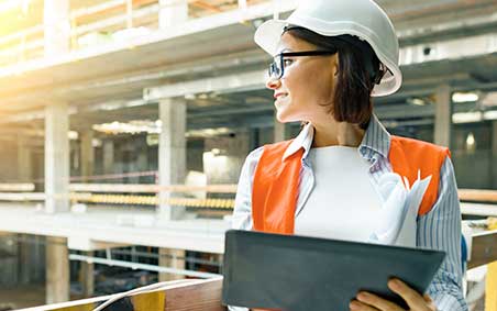 A woman in a construction site wearing a hard-hat, safety glasses and a safety vest, working on a tablet.