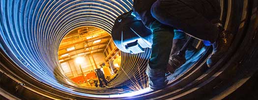 Welder standing inside of a large corrugated metal pipe performing a weld on the interior of the pipe underneath.