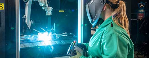 A female welding operator wearing a RADNOR welding helmet, operates an automated welding machine performing a weld.  