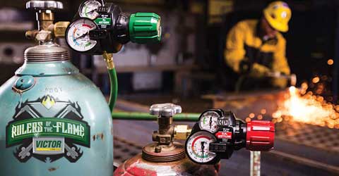 Two gas cylinders featuring Victor gas regulators are positioned in the foreground to the left, while in the background to the center-right and slightly out of focus, a welder uses a Victor cutting torch.