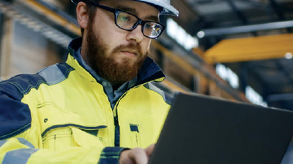 A man wearing personal protective equipment (PPE) in an industrial plant, warehouse or facility, types on a laptop computer.