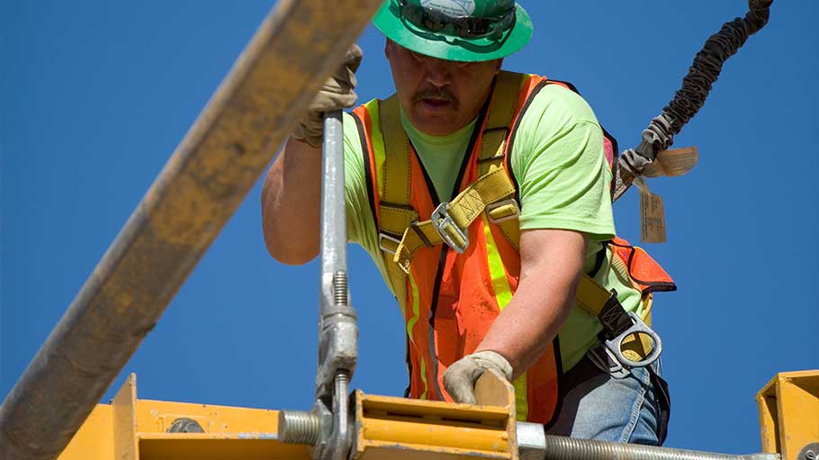 A construction worker wearing PPE, including fall protection, adjusting a bolt with a large wrench