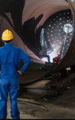 A man wearing a hardhat and protective overalls watching as a welder welds inside a large pipe.