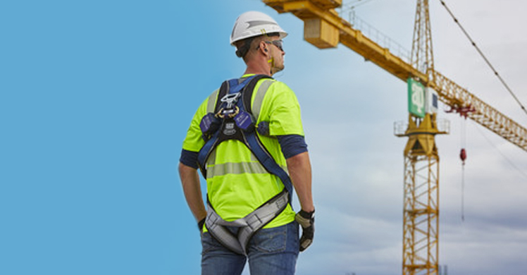 Back view of construction worker wearing safety harness, neon jersey & white hardhat against blue sky, crane in upper corner.