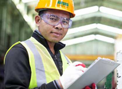 Set in a warehouse or industrial plant or facility, a man wearing personal protective equipment (PPE), including an Airgas branded hardhat, high-visibility safety vest and safety gloves, holds a clipboard.