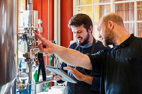 Two brewers checking gas levels in a fermentation tank or brite tank while referencing notes on a tablet.