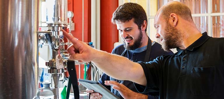 Two brewers checking gas levels in a fermentation tank or brite tank while referencing notes on a tablet.