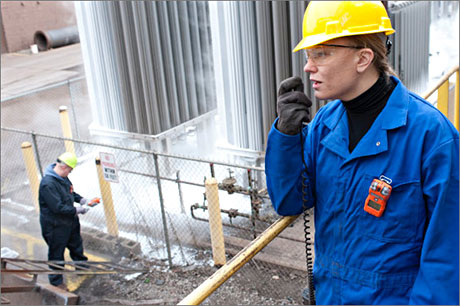 Woman on-site talking into 2-way radio wearing a hardhat, protective gloves, and a gas monitor clipped on the chest pocket of her coverall.