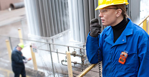 Woman on-site talking into 2-way radio wearing a hardhat, protective gloves, and a gas monitor clipped on the chest pocket of her coverall.