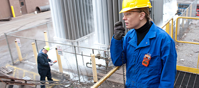 Woman on-site talking into 2-way radio wearing a hardhat, protective gloves, and a gas monitor clipped on the chest pocket of her coverall.